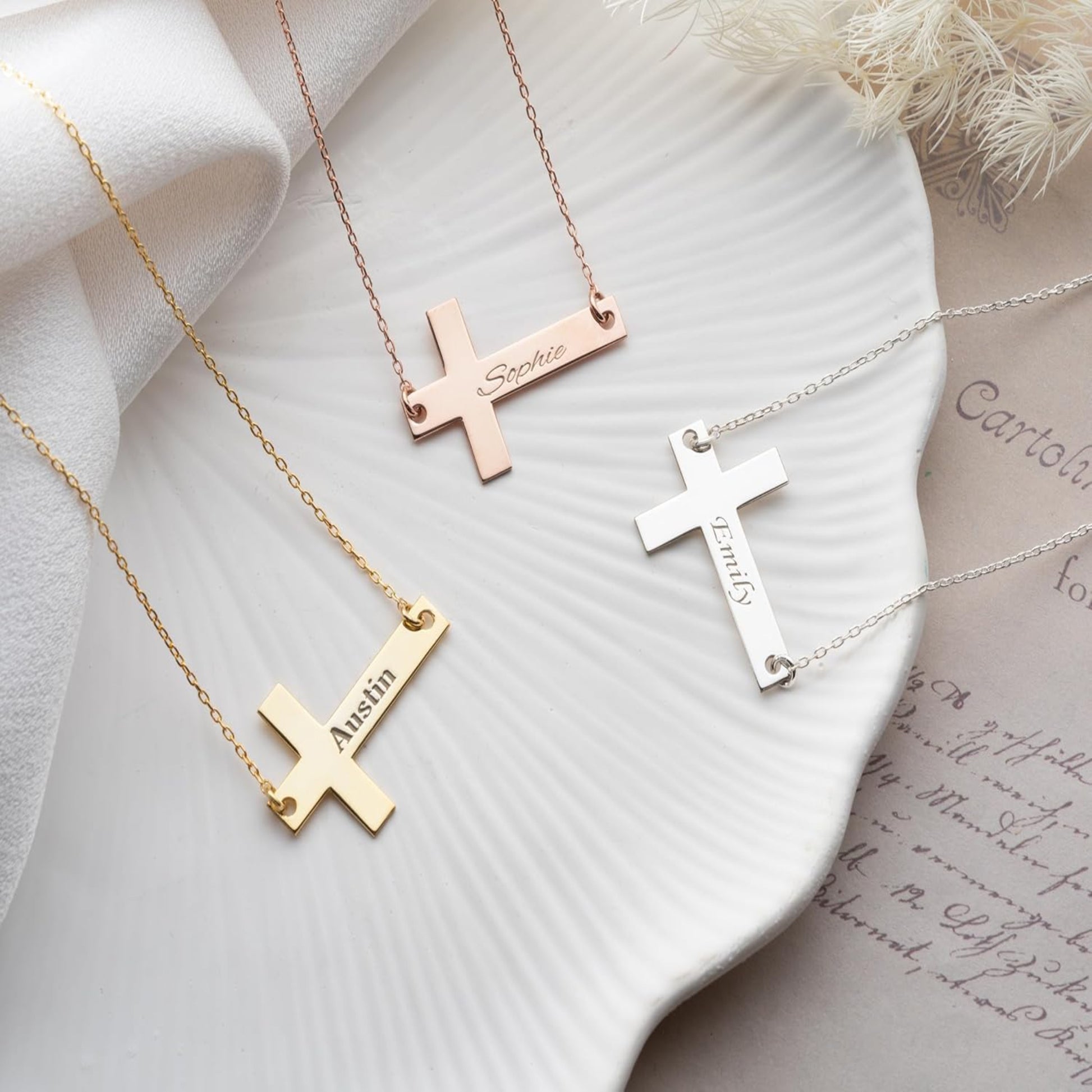 Three cross-shaped necklaces with engraved names on a textured white surface.