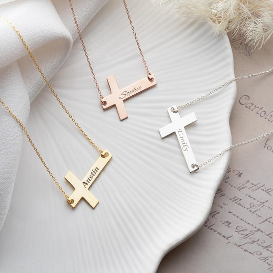 Three cross-shaped necklaces with engraved names on a textured white surface.