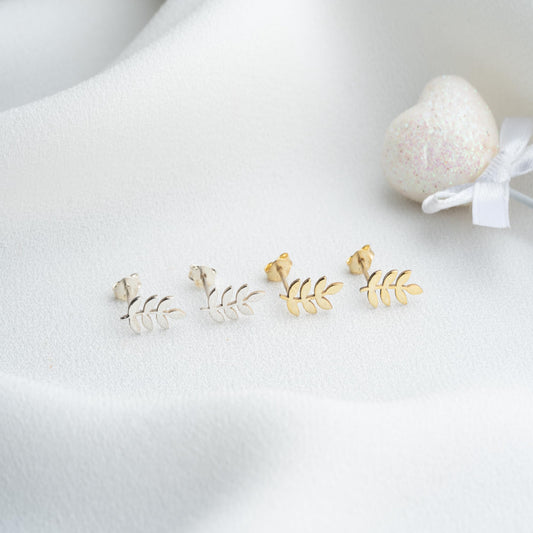 Gold leaf-shaped earrings on a white surface with a heart-shaped object in the background.