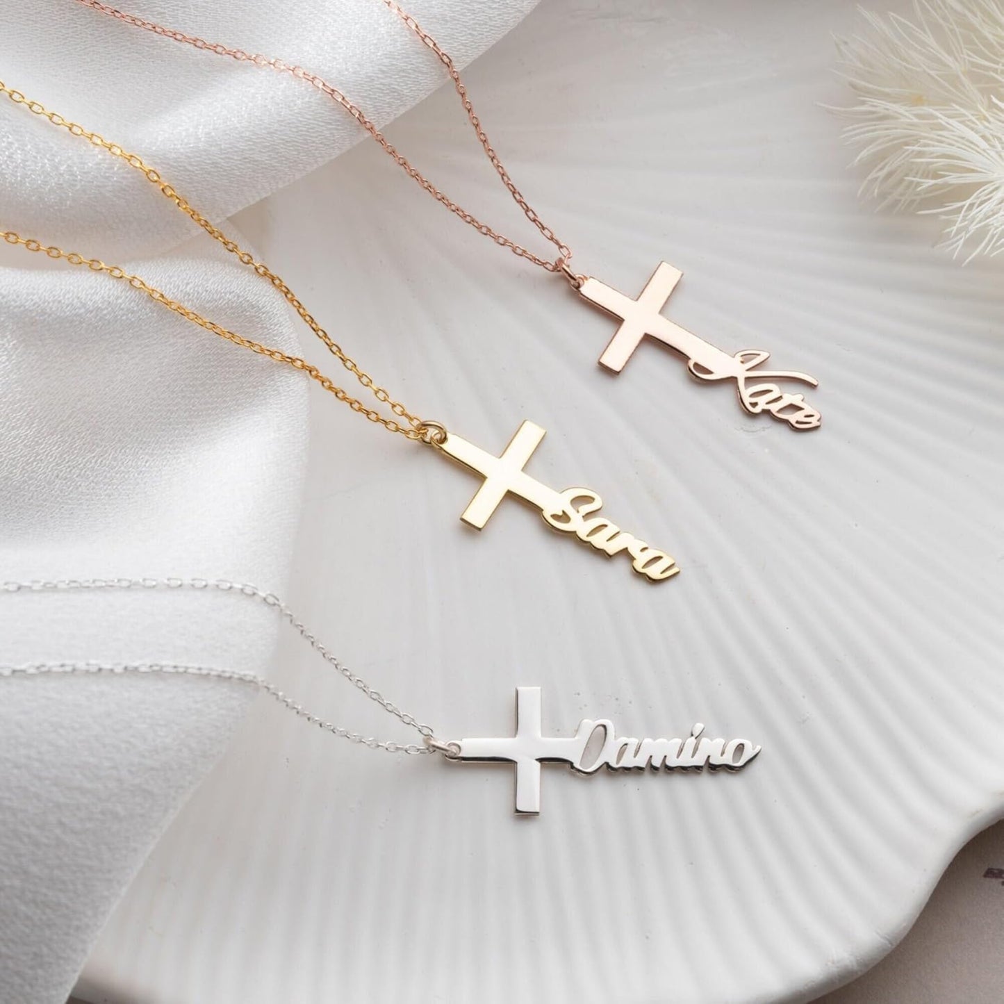Three gold cross-shaped necklaces with engraved names on a white fabric background