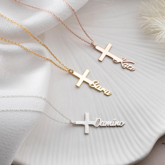 Three gold cross-shaped necklaces with engraved names on a white fabric background